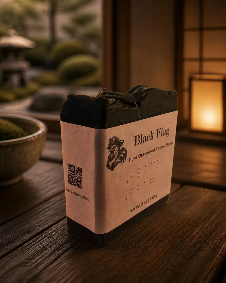 Black Flag tallow soap with Brailled paper label on warm wood surface, lantern glow in background.