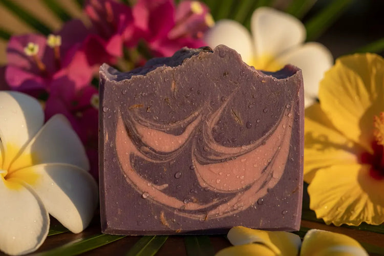 Close-up of rich purple Alani Moani soap bar with pink rose clay swirls and water droplets, tropical island flowers in background