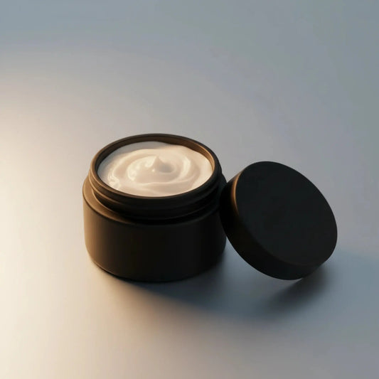 An open matte black cosmetic jar filled with white cream, with its matching lid leaning against it on a clean, light gray background.