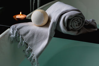 Large bath bomb on soft blue cotton towel with lit candle on bathtub tray in dimly lit bathroom creating spa feel.