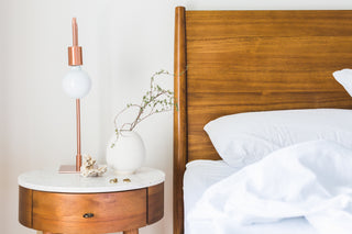 Minimalist wooden bedside table with lamp, plant, and seashell earrings beside bed with white linens