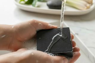 Luxurious lather from charcoal tea tree black soap bar in hands with tea tree leaves and lemongrass in spa background