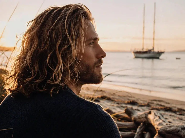Man with long wavy tallow-washed hair on beach at golden hour, sailboat anchored offshore, Blind Pirate Soap Co.