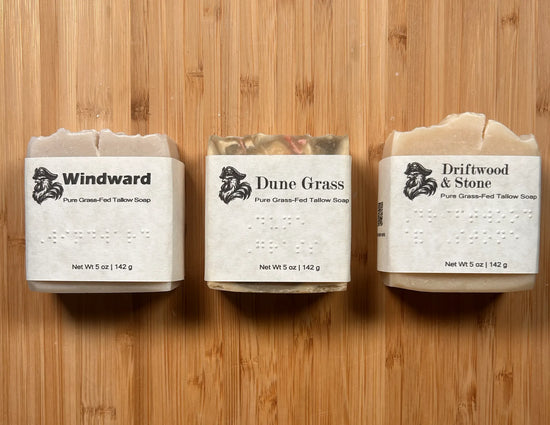 Windward, Dune Grass, and Driftwood & Stone — three wrapped Blind Pirate tallow soap bars side by side on bamboo, Braille labels visible
