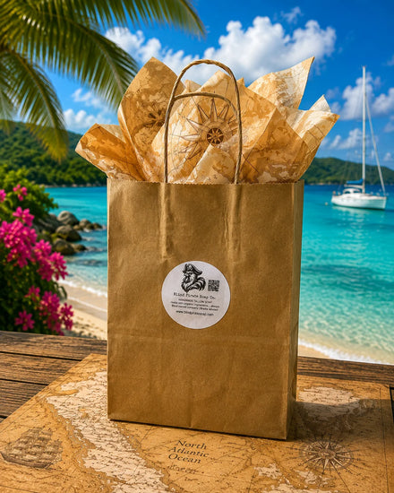 Blind Pirate Soap Co. Stowaway Discovery Set kraft gift bag with tissue paper on a tropical beach, turquoise water and sailboat behind
