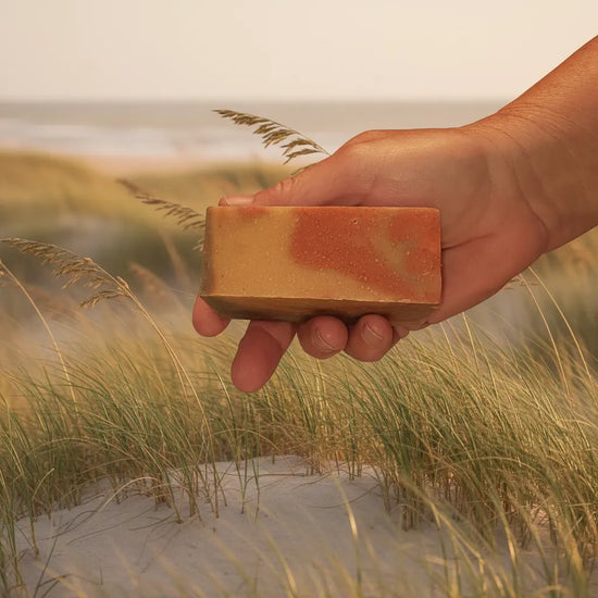 Dune Grass artisan soap bar by Blind Pirate Soap Company — warm terracotta and golden swirl, held against a sandy coastal dune scene.