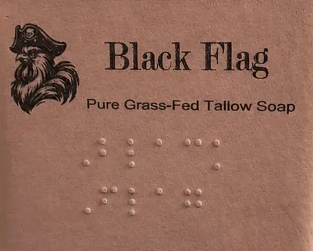 Black Flag label close-up — pirate logo, title, and full Braille block, warm lantern glow behind. Blind Pirate Soap Co.
