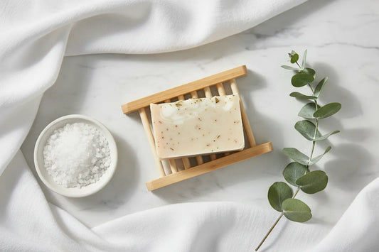 Flat lay of handmade artisan soap bar on bamboo tray on white marble surface surrounded by white linen, sea salt crystals, and fresh eucalyptus sprig