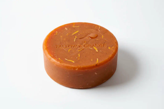 shampoo bar orange and honey, organic oil shampoo bar sits on a white back ground. The bar has been stamped with the words tallow and orange on the top and it has flecks of a lighter yellow and darker orange throughout. The bar is puck sized and round.