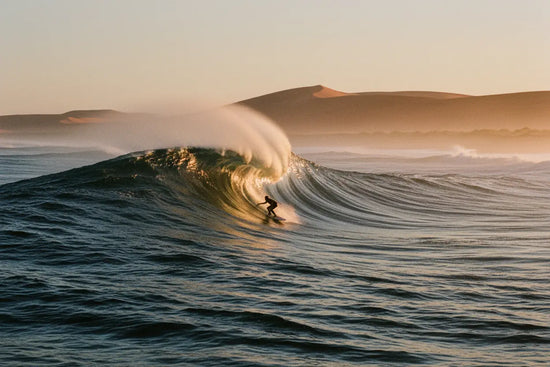 Surfer Suds | A lone surfer threading the barrel at Skeleton Bay, Namibia — dawn light electric on the wave's glassy wall.