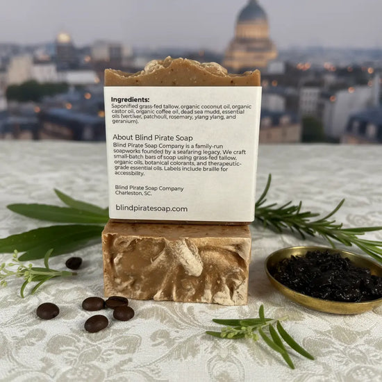 Back panel of Stone Ground tallow soap bar beside caviar and roasted coffee beans, golden Paris skyline glowing softly in the distance.