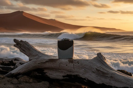 Surfer Suds unwrapped tallow soap bar on sun-bleached driftwood, wild Skeleton Bay, Namibia stretching beyond.