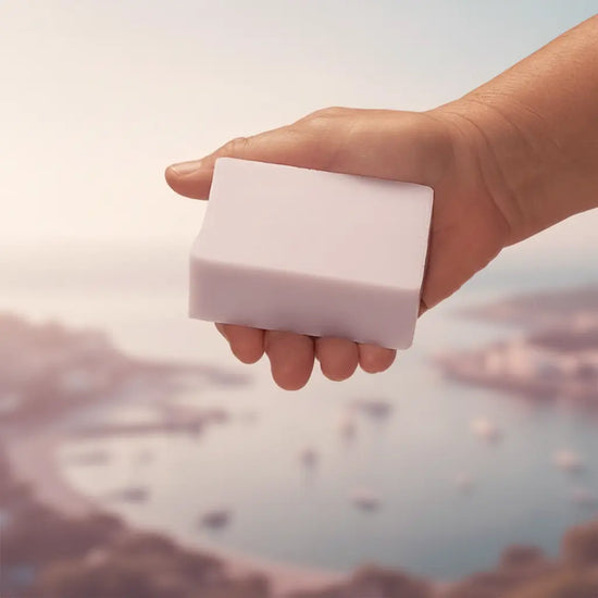 Windward tallow soap bar held in hand above a soft mauve coastal seascape, warm hazy light. Blind Pirate Soap Co.
